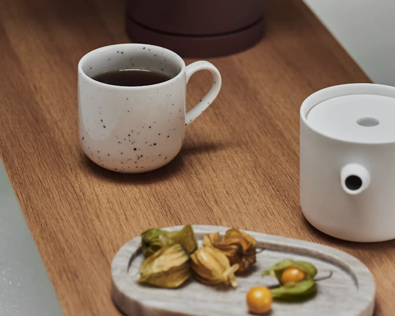 斑点模様の白いマグカップに濃い飲み物、蓋付きの白い容器、木製トレイにホオズキ、全て木製の表面に。